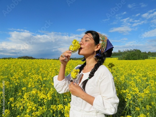 Принти на полотні Woman in white dress in yellow canola field holding wine glass with flowers