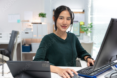 Smiling female customer service representative wearing a headset while working at a desktop computer in a modern office. Confident support agent looking at the camera, providing online customer servic
