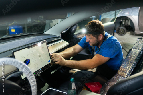 Car cabin shows touchscreen and open glove box in workshop bay. Technician taps icons testing settings with flashlight during diagnostics