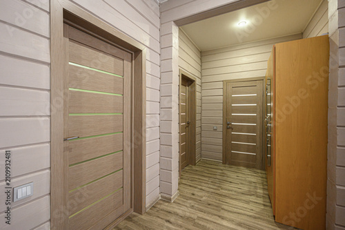Canvastavla narrow hallway in wooden house: light‑toned wood‑effect panel walls, beige wood‑grain doors with horizontal inlays, a tall warm‑toned wardrobe, wood‑look flooring under recessed ceiling lighting