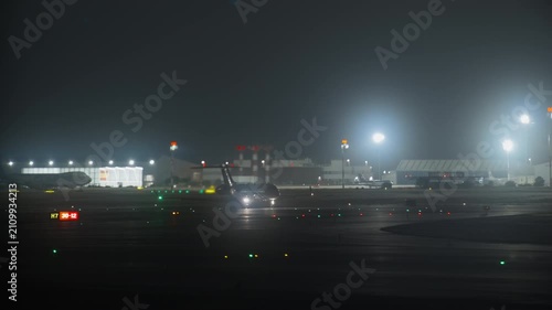 Narrow body aircraft taxiing on night apron, tilt shift lens effect creating selective focus, colorful ramp light and bokeh, terminal and hangar illuminated in background