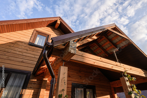 wooden house features warm-toned cladding, gabled roof with exposed rafters, dark metal gutters, stone supports, and climbing vines under blue sky with wispy clouds