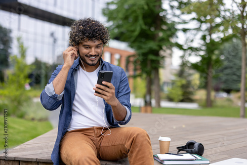 Bild auf Leinwand Smiling young man with curly hair sitting outdoors on a university campus and pu