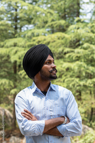 Obraz na plátně Vertical portrait of a young Sikh man wearing a black turban and a blue shirt, standing with arms crossed while looking away against a blurred background of lush green trees in Himachal Pradesh