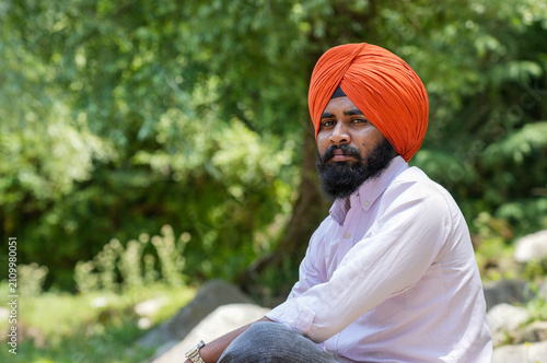 Foto Closeup portrait of a young Sikh man wearing an orange turban, looking directly at the camera while sitting against a blurred background of lush green trees in Himachal Pradesh, with empty copy space