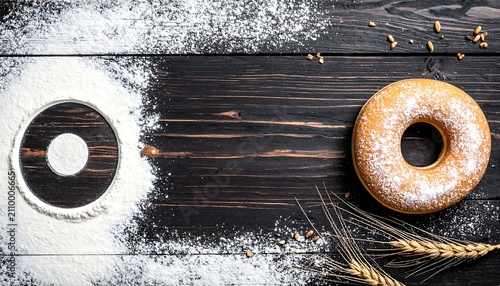Top view of fresh donut with powdered sugar, flour, and wheat ears on dark wooden background with copy space