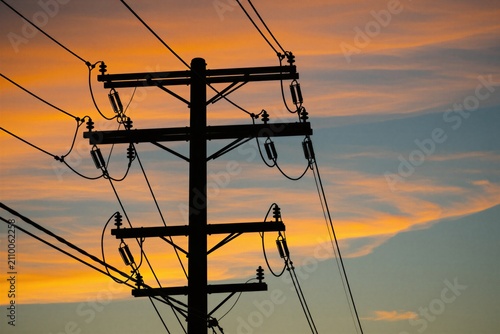 Silhouette of electrical utility pole against colorful orange sunset sky. High voltage wires, metal cables stretch across horizon during twilight. Urban industrial infrastructure stands dark against