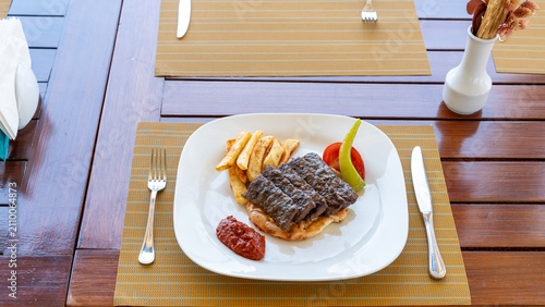 Fotografie Hearty Meal Presentation: A plate of grilled meat served with french fries, a slice of tomato, and a dollop of ketchup, set on a wooden table, promising a satisfying dining experience