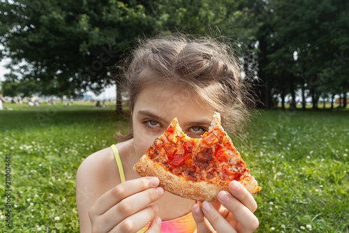 Cute young girl holding a bitten slice of pizza in nature park