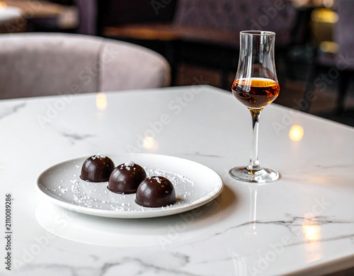 A plate of dark chocolate pralines and a small glass of liqueur sit on a marble-topped bistro table.