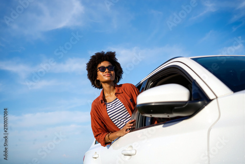 A woman with curly hair and sunglasses leans out of a white car, soaking in the warm sun and fresh air while gazing at the beautiful sky during the day. Generative AI.
