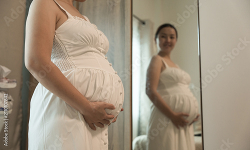 Close-up of a pregnant woman's belly in a white dress, with her smiling reflection visible in the mirror behind.