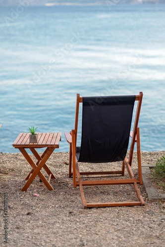 Wooden deck chair with black fabric and small table beside it overlooking calm blue water
