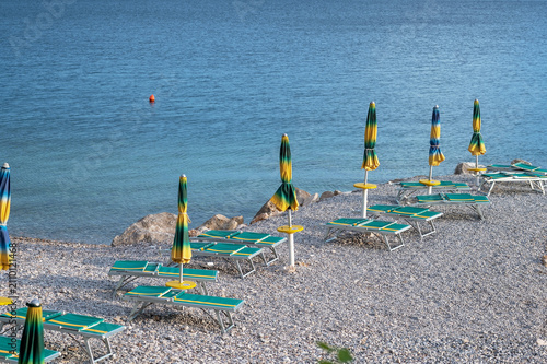 Beachside lounge chairs with colorful umbrellas arranged on pebbled shore near calm blue water