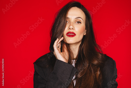Sensual glamorous young woman touching hair face looking at camera parted lips direct gaze wearing dark blazer red lipstick, copy space isolated on studio bright red background beauty stock photo