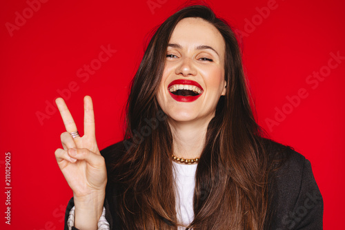 Joyful laughing young woman showing peace sign victory gesture looking at camera broad smile teeth open mouth wearing dark blazer red lipstick, isolated on studio bright red background photo