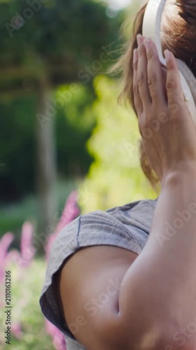 Vertical video: Tapping right earcup grey top woman fitting phones for music in garden, copy space