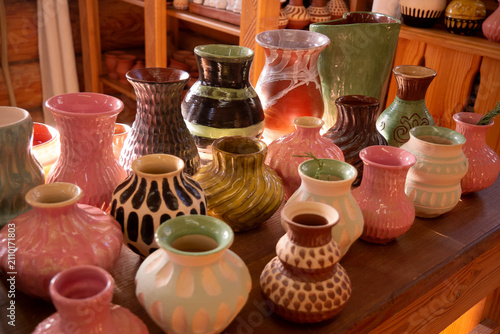 A collection of colorful handmade ceramic vases on a wooden table, viewed from above. The vases feature a variety of shapes, textures, and earthy tones, and are finished with a glossy glaze