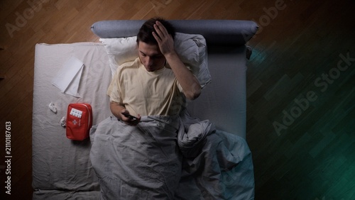 Canvas Print Young unwell man lying in bed at home, experiencing a headache and looking at hi