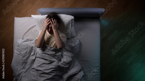 Fotografía Young man lying in bed late at night, covering his face with hands, experiencing