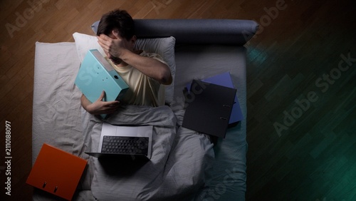 Fotografie Man lying in bed covering his eyes with his hand, surrounded by files and a lapt