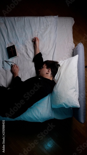 Obraz na plátně Priest resting in bed, wearing a cassock and holding a rosary and bible, symboli