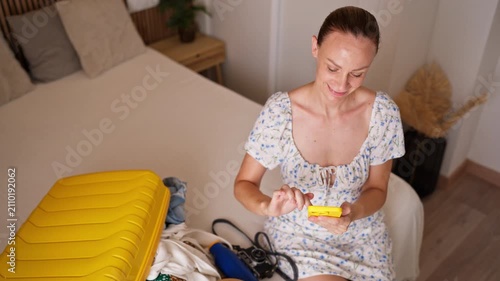 Young woman booking travel online using smartphone while sitting near open yellow suitcase in bedroom, smiling and getting ready for vacation trip