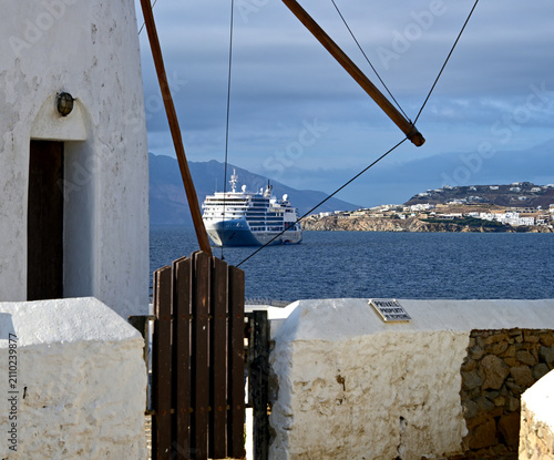A large cruise ship anchored in the bay off Mykonos, Greece, framed by traditional whitewashed architecture and rustic wooden details. The scene captures the contrast between classic Cycladic design a
