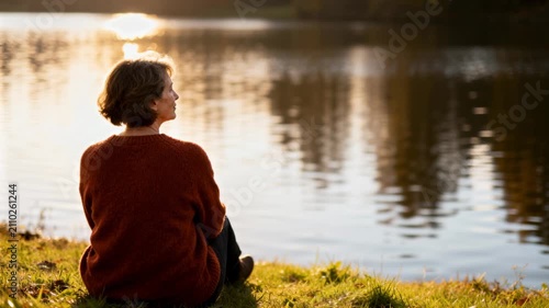 Senior woman sitting on grass by a lake at sunset. Back view of a female reflecting by the water during golden hour in autumn. Retirement and solitude concept
