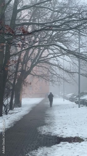 Foggy park walkway with lone figure. Bare branches arch over brick path with patches of snow, distant lamp posts and building silhouette, Taken on mobile device.