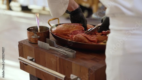Closeup of gloved chef carving roasted duck with golden potatoes in copper pan. Warm lighting highlights rich brown golden tones. Rustic wooden cart with dark ceramic plates ready for serving