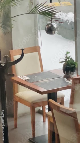Cozy cafe table with chairs and plants, wooden table near large window under pendant lamp, potted greenery and calm morning ambience Taken on mobile device.