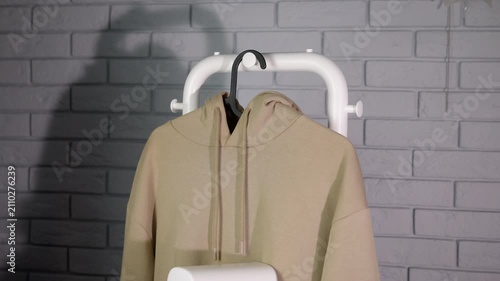 Woman steams clothes. Person steaming a beige hoodie with a portable iron steamer against a gray brick wall, performing household chores