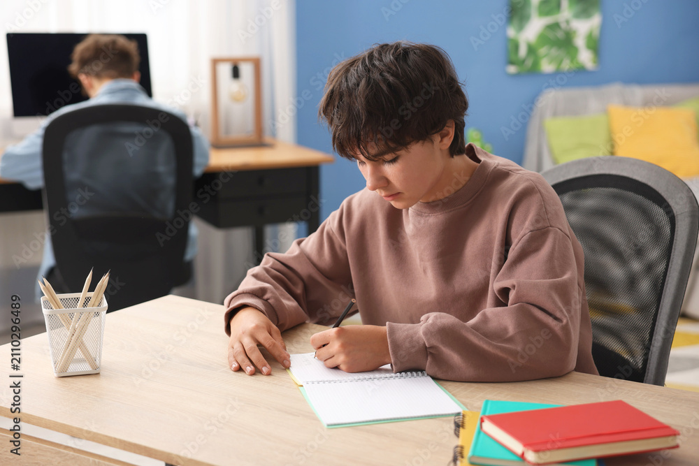Naklejka premium Teenage boy doing homework at wooden table while his brother sitting with computer at home, selective focus