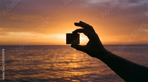 Silhouette of Human Hand Holding Small Wooden Cube Block Against Vibrant Orange Sunset Sky Over Calm Ocean Horizon, Golden Hour Light Reflection on Water Surface, Minimalist Abstract Nature