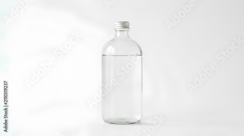A pristine clear glass bottle filled with pure water stands isolated against a seamless white background, featuring a silver screw cap and soft studio lighting perfect for product mockups.
