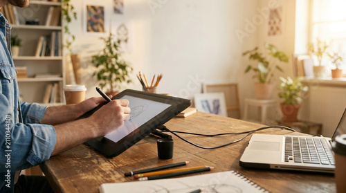 Professional graphic designer illustrator working on digital drawing tablet with stylus pen connected to laptop computer in cozy sunlit home office studio filled with green house plants.