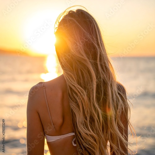 A woman with long blond hair facing away from the camera, at sunset. A bright orange glow from the sun is visible