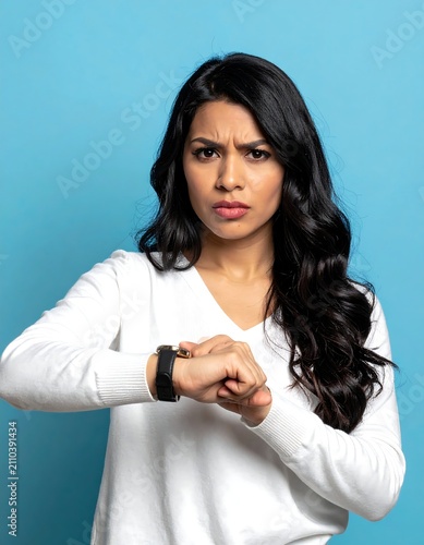 A woman with long dark hair, wearing a white sweater, and a smartwatch, looks at it with an angry expression against a blue backdrop