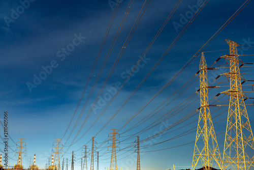 High voltage electricity transmission towers and power lines under a blue night sky with stars near an electrical substation. Modern energy infrastructure, utility networks and power distribution.