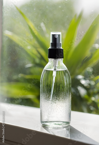 A bottle of water sitting on a window sie, close up of a minimalist clear glass spray bottle with fine water droplets misting gently into soft window light
