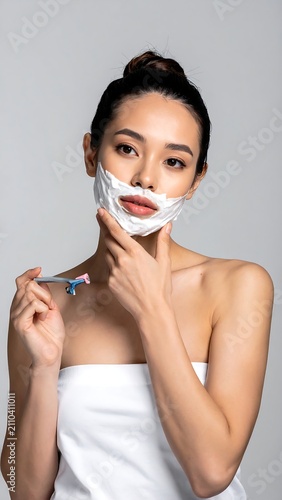 A young, Asian woman with a towel wrapped around her torso shaves her face. Her hair is up, and shaving cream covers her lower face