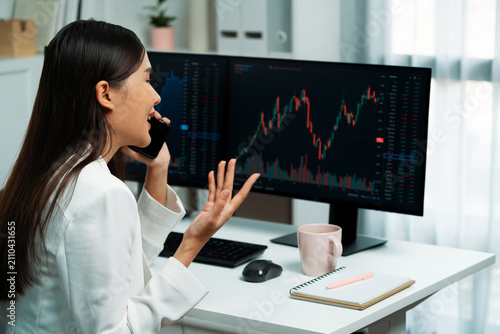 Fotografía Smiling young beautiful Asian businesswoman calling to broker on smartphone to analyze exchange stock market graph on pc screen at modern office