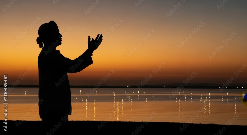 Fototapeta premium Silhouette of a person praying with hands raised at sunset by the calm sea
