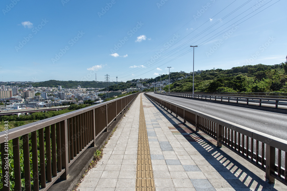 Fototapeta premium 快晴の空と左側と正面の斜面に住宅地のある橋の点字ブロックが続く歩道と車道