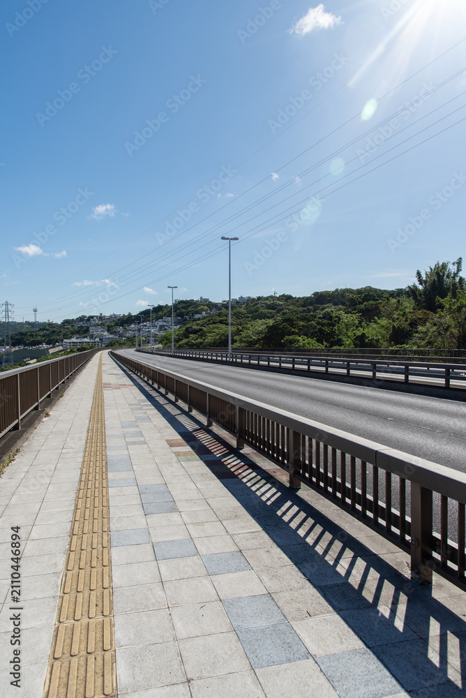 Fototapeta premium 快晴の空と正面の斜面に住宅地のある橋の点字ブロックが続く歩道と車道