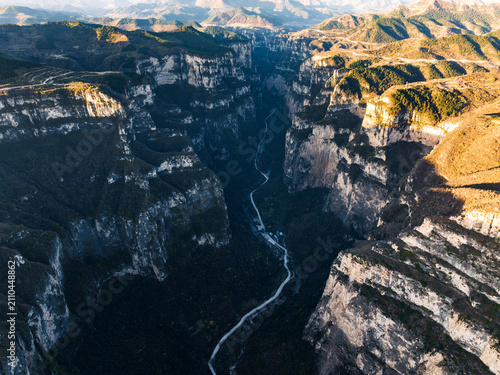 A deep, narrow canyon with steep rocky cliffs, a winding road running along its floor, under sunlight with distant mountain ranges.