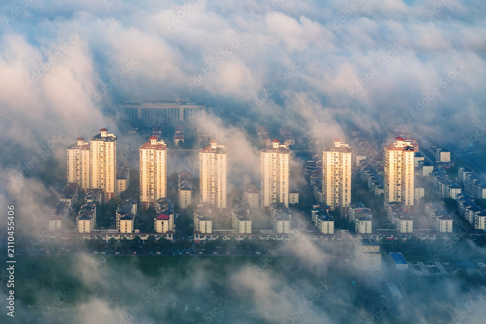 Fototapeta premium Aerial view of high-rise residential buildings partially shrouded in dense morning mist, with warm daylight illuminating their facades.