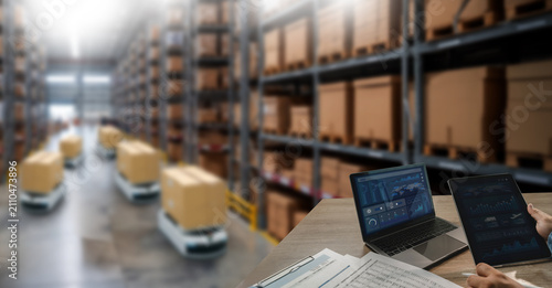 Warehouse robots transport boxes through storage aisles while a laptop and tablet display operational dashboards, supporting logistics automation, inventory monitoring, and warehouse management.