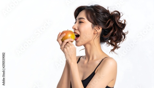 A young woman with dark hair tied back, smiles broadly while taking a bite of a ripe, juicy apple. She is dressed in a simple black top